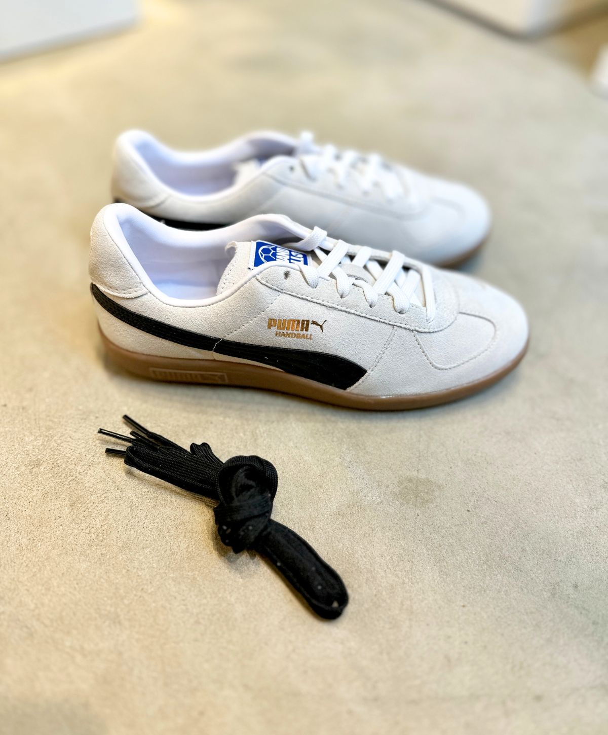 Et par hvide Puma håndboldsneakers med sorte detaljer og en brun gummisål, placeret på en lys overflade. Ved siden af skoene ligger et sort skosnøre.