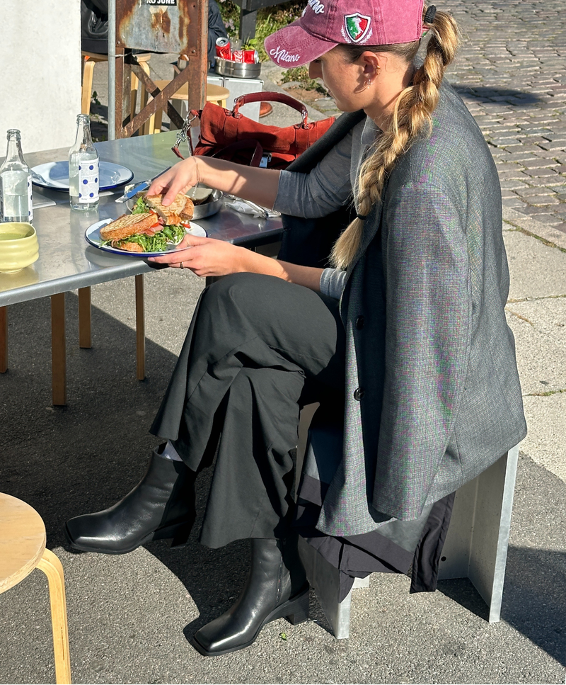 En person sidder ved et udendørs bord og spiser en sandwich. Personen har en grå jakke, sorte bukser og sorte støvler på. Der er en lyserød kasket på hovedet. På bordet er der en flaske og en skål.