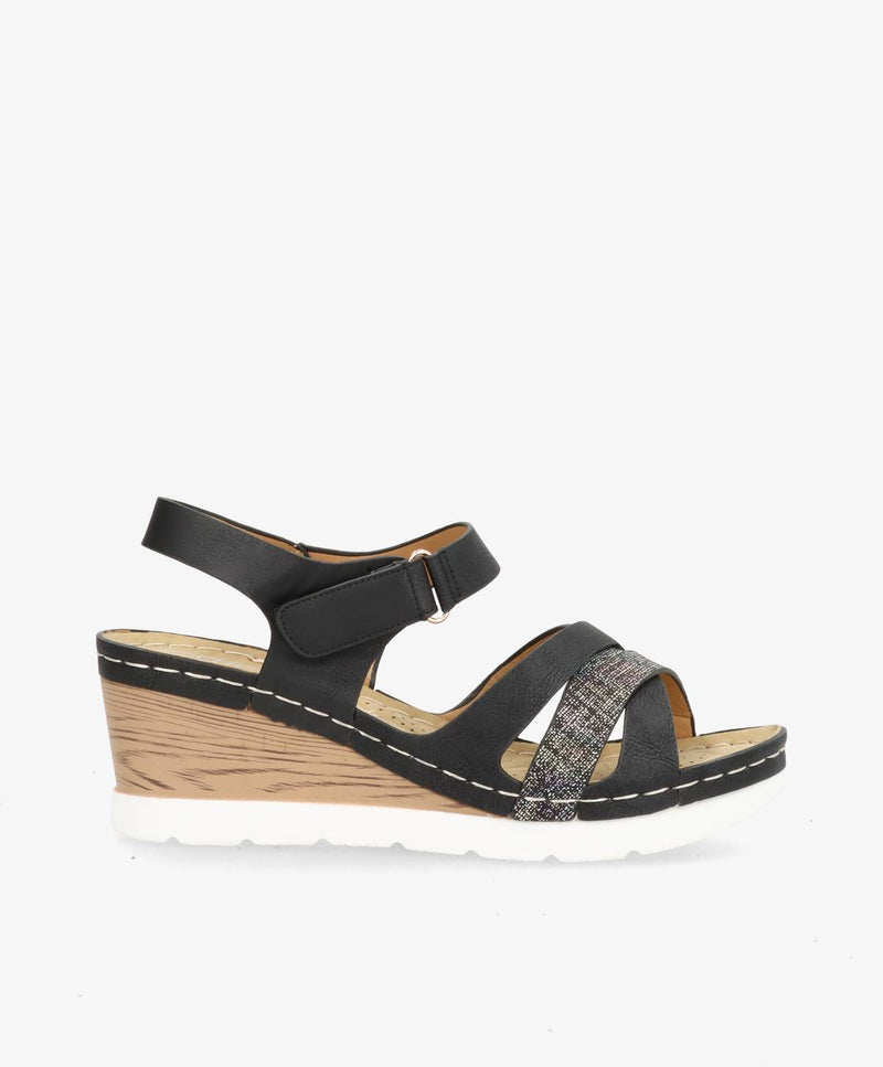 Sort sandal med kilehæl, justerbar rem og dekorative detaljer på stropperne.