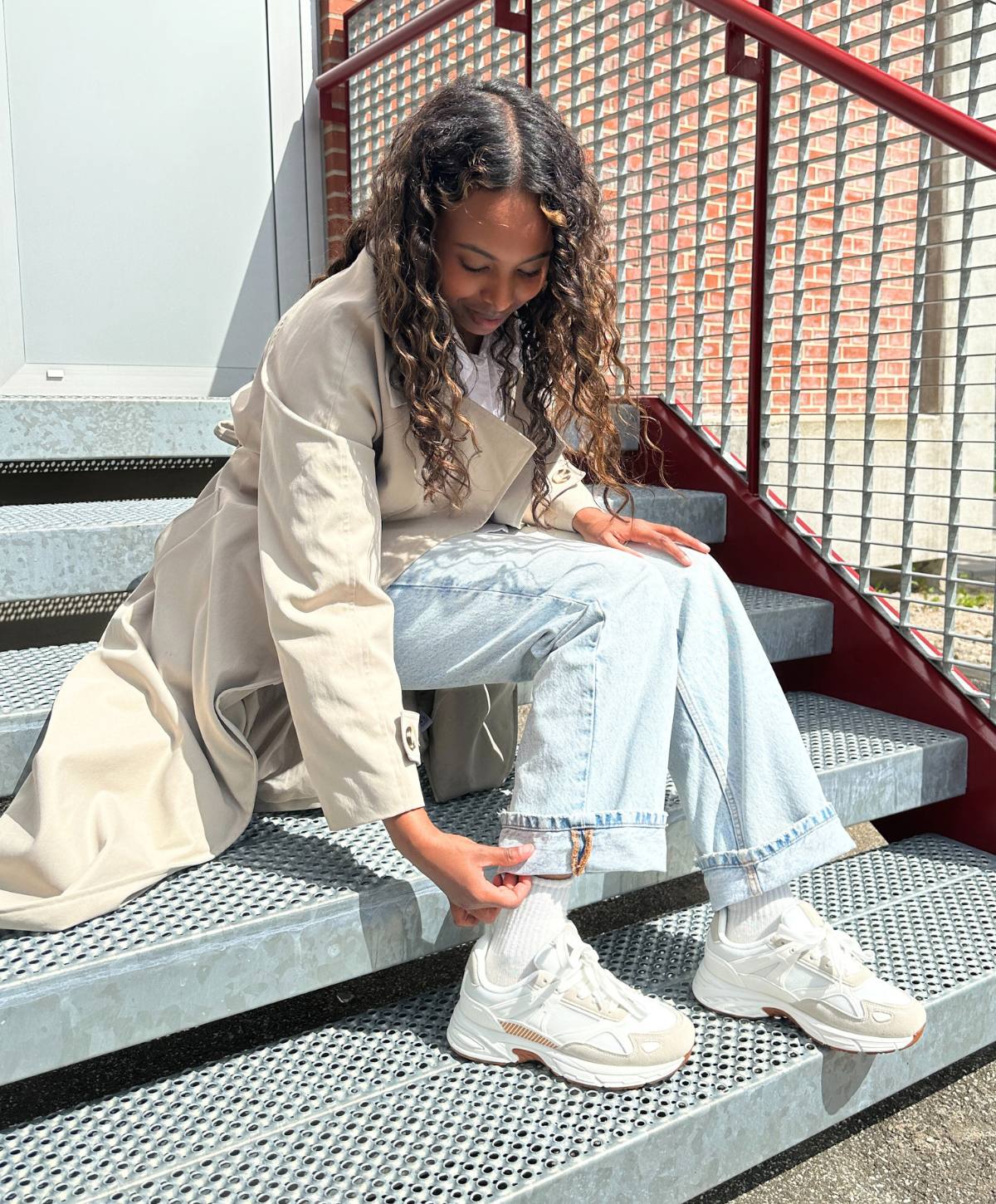 En kvinde med langt, krøllet hår sidder på en metaltrappe iført en beige frakke, lyse jeans og hvide sneakers. Hun justerer kanten af sine jeans.