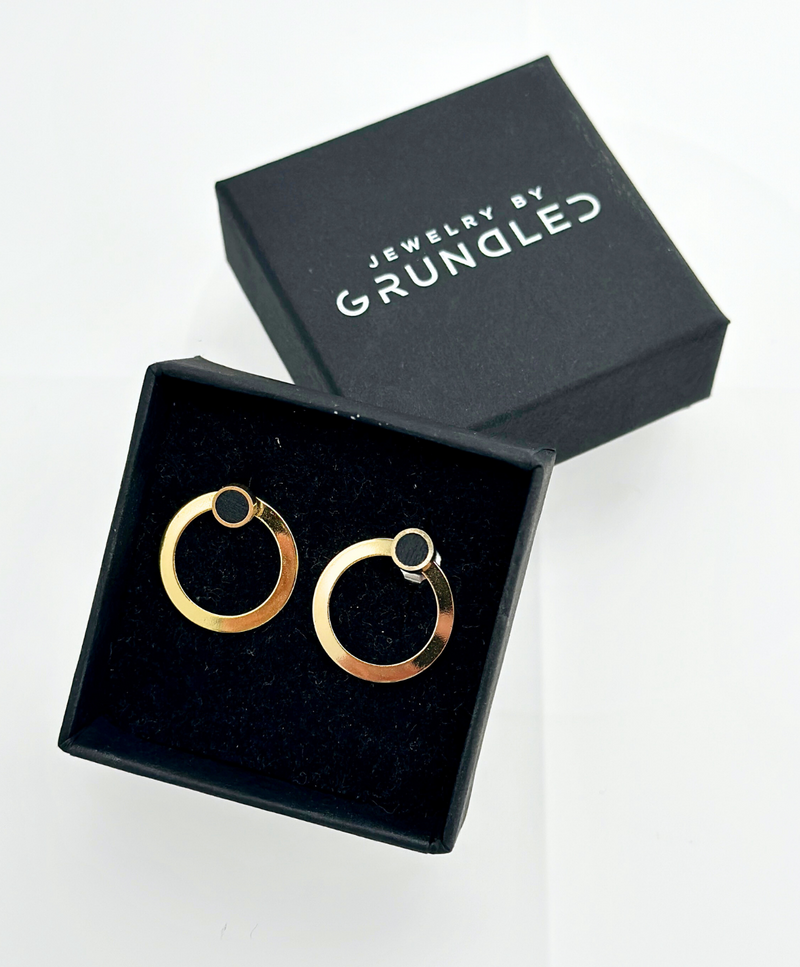 Sort smykkeæske med teksten "Jewelry by Grundled" indeholder et par guldøreringe med cirkulært design.