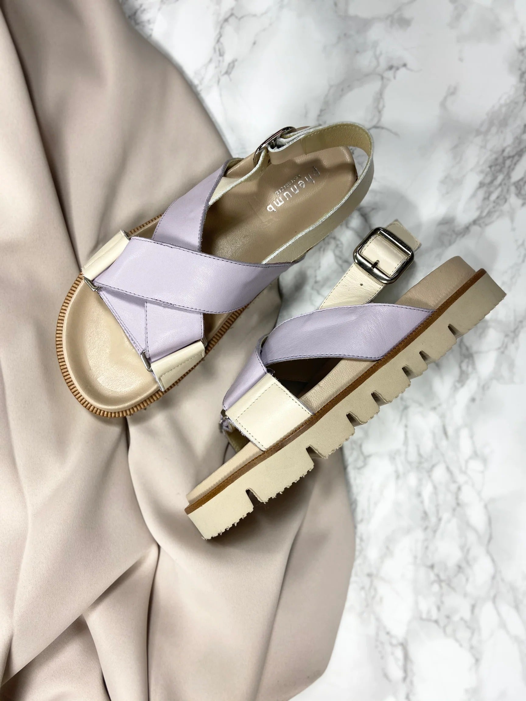 Sandaler med krydsede stropper i lys lilla og beige læder på en marmorbordplade.